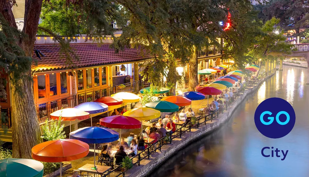 An outdoor riverside dining area illuminated at night with vibrant colorful umbrellas and people enjoying their evening