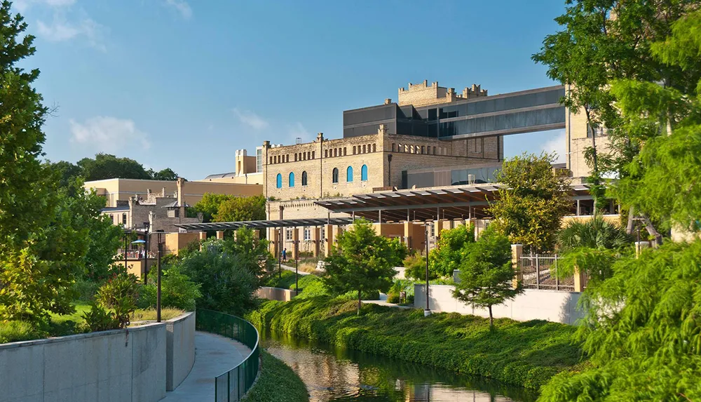 The image depicts a modern urban park with a walkway along a river lush greenery and contemporary buildings in the background