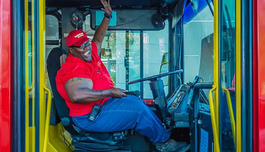 A smiling bus driver in a red shirt and cap is seated at the wheel of a bus, waving to the camera with a friendly gesture.