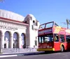 A red double-decker sightseeing bus is parked in front of the historic Alamo mission under a clear blue sky