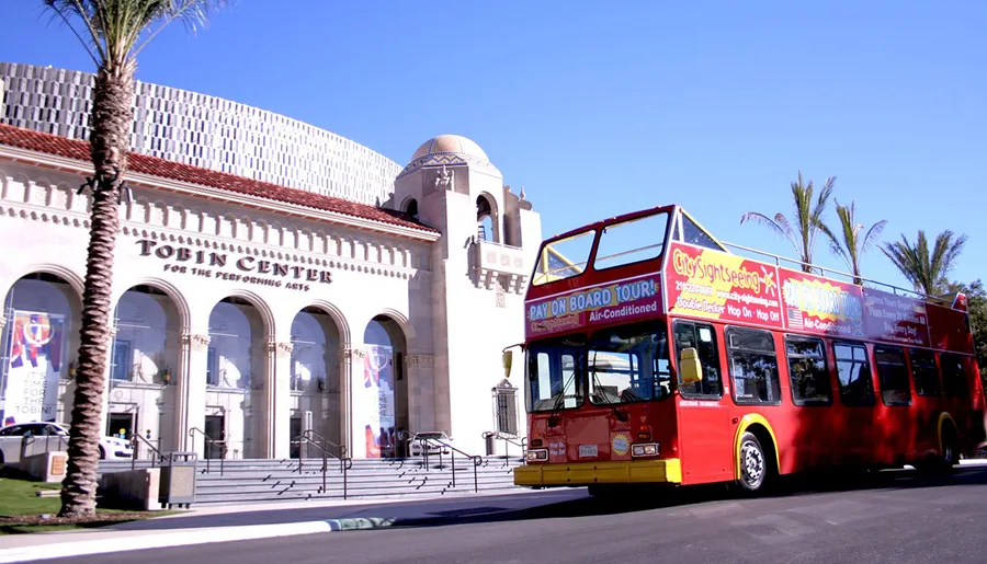 A red double-decker sightseeing tour bus passes in front of the Tobin Center for the Performing Arts under a clear blue sky.