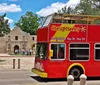 A red double-decker sightseeing bus is parked in front of the historic Alamo mission under a clear blue sky