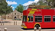 A red double-decker sightseeing bus is parked in front of the historic Alamo mission under a clear blue sky.