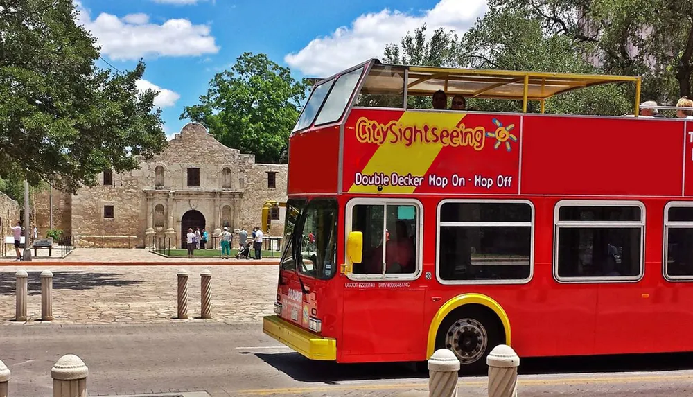 A red double-decker sightseeing bus is parked in front of the historic Alamo mission under a clear blue sky