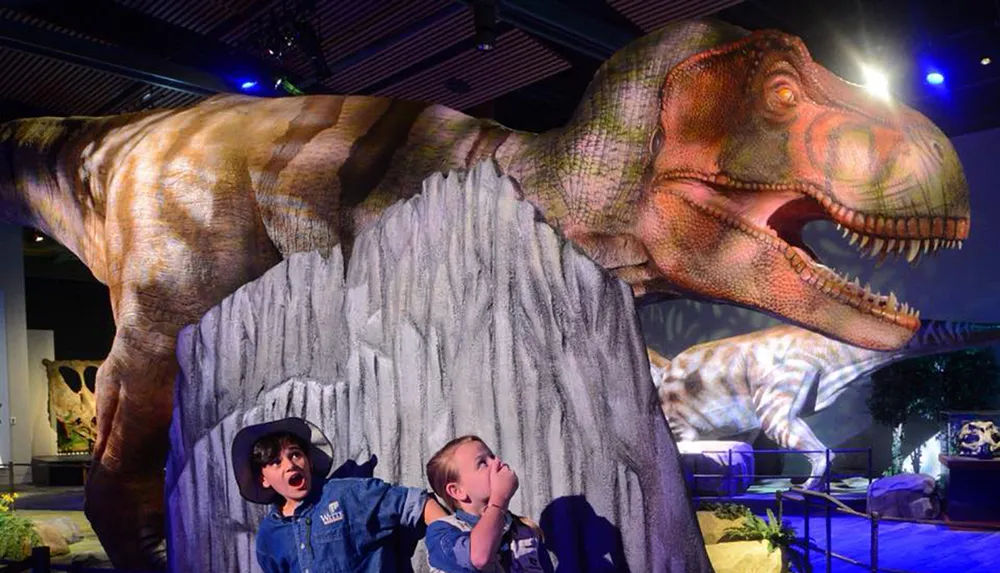 Two children are playfully reacting to a large dinosaur exhibit giving the impression of a thrilling prehistoric encounter