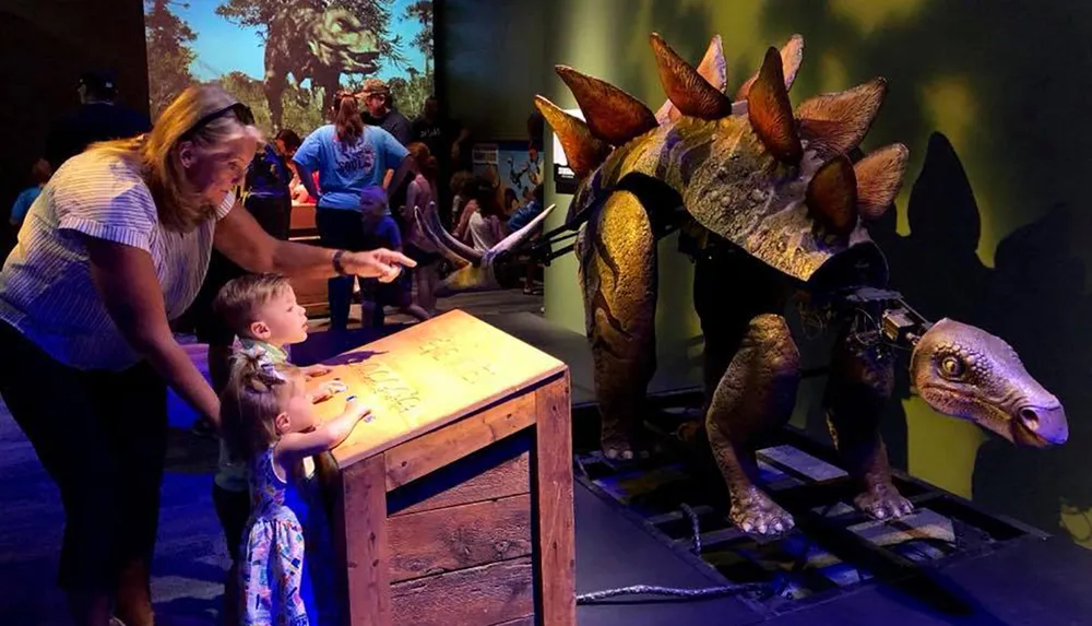 An adult and two young children are looking at what appears to be a life-size animatronic dinosaur in an exhibition
