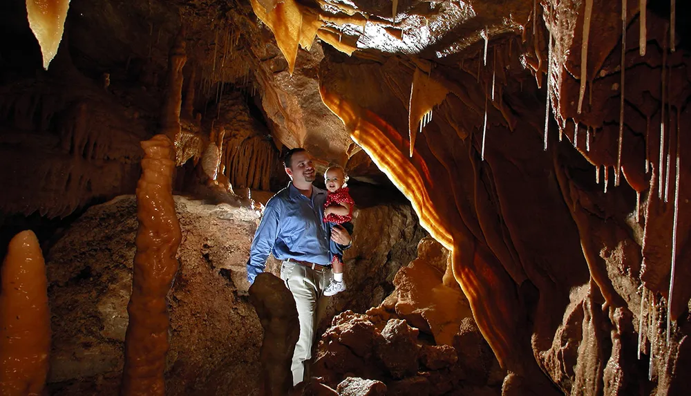 A smiling person is holding a happy child inside a richly textured cave lit warmly highlighting the caves stalactites and stalagmites