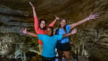 Three joyful individuals are posing with arms raised in a cave-like setting, expressing happiness or excitement.