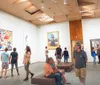 A person admires artwork in a gallery while another takes a photo with their smartphone