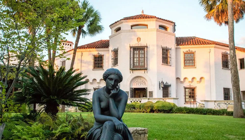 The image shows a bronze statue of a seated contemplative woman in front of an elegant white stucco building with Spanish architectural features set amid lush greenery and palm trees in what appears to be a tranquil garden setting