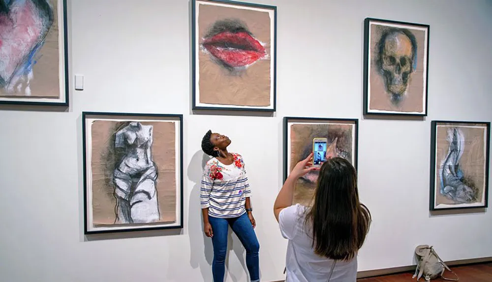 A person admires artwork in a gallery while another takes a photo with their smartphone