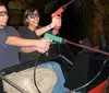 Two people are smiling and wearing 3D glasses seemingly enjoying a ride or attraction where they hold a toy gun together against a backdrop that resembles a stone tunnel