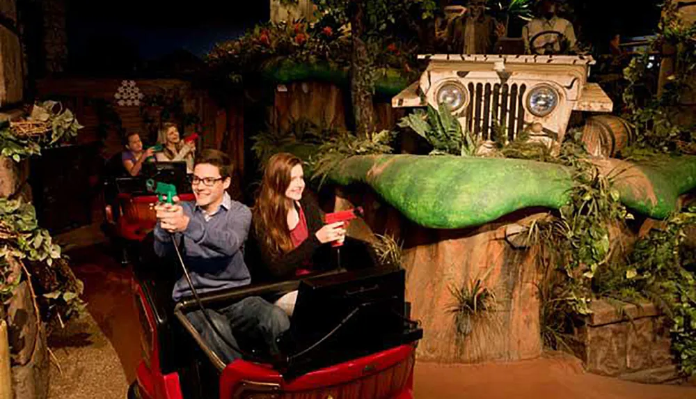Visitors are enjoying an indoor interactive ride where they are seated in a vehicle designed to look like a red jeep and are holding plastic guns presumably to shoot at targets within a jungle-themed set that includes a life-sized model of a jeep