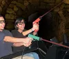 Two people are smiling and wearing 3D glasses seemingly enjoying a ride or attraction where they hold a toy gun together against a backdrop that resembles a stone tunnel