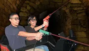 Two people are smiling and wearing 3D glasses, seemingly enjoying a ride or attraction where they hold a toy gun together, against a backdrop that resembles a stone tunnel.
