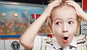 A surprised young child is wide-eyed and holding their head with their mouth open in front of a backdrop featuring the Guinness World Records logo.