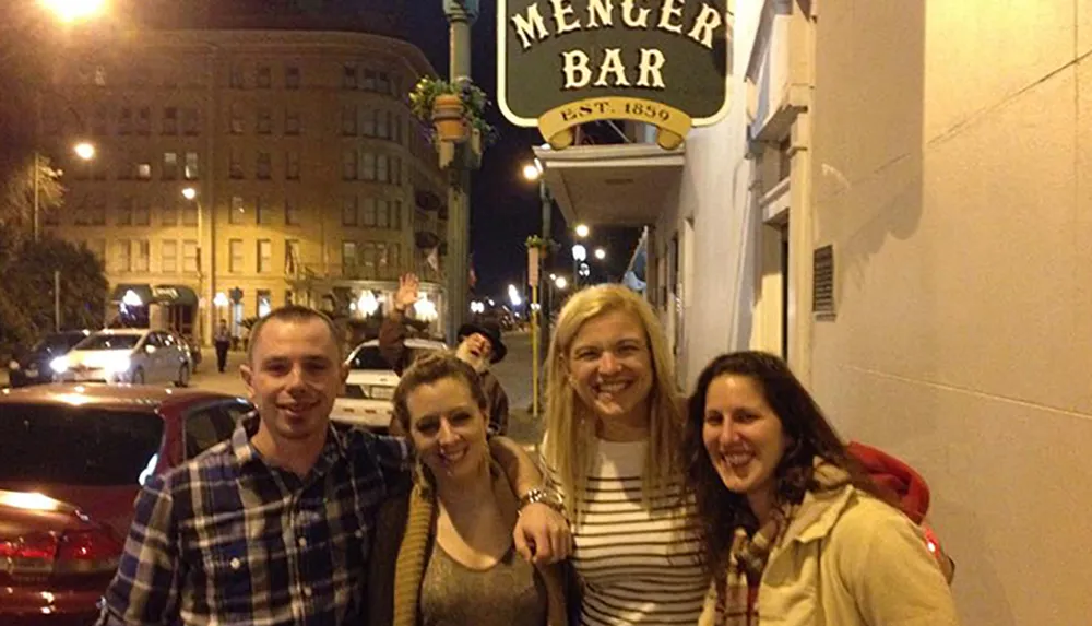 Four people are posing for a photo while smiling on a city street at night in front of the Menger Bar with someone photo-bombing in the background
