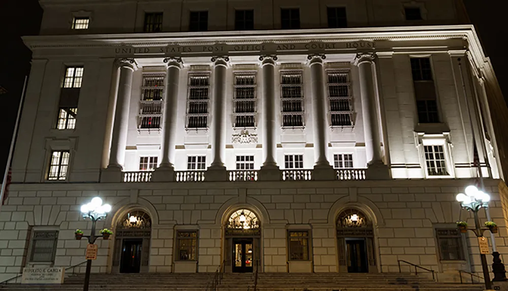 The image shows the illuminated facade of the United States Post Office and Court House at night exhibiting neoclassical architectural columns and a grand staircase entrance