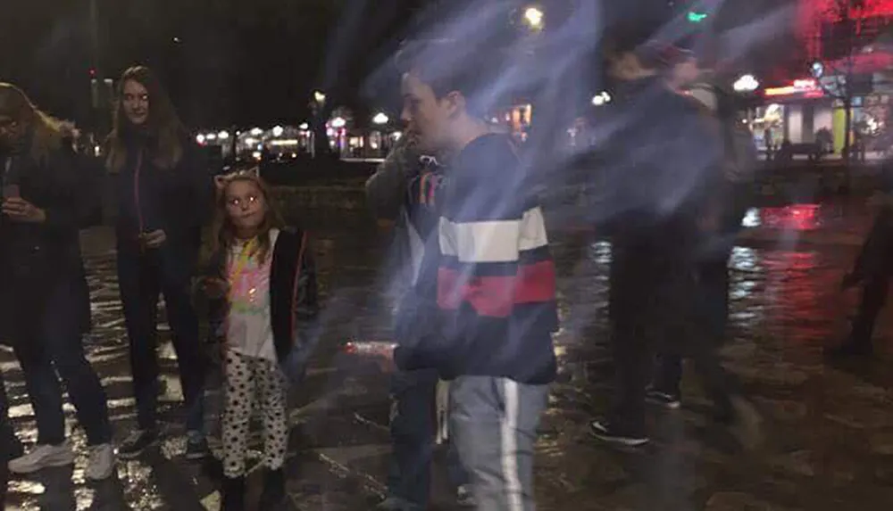 A group of individuals appears to be standing on a wet street at night with some motion blur suggesting movement and various bright light sources in the background creating a lively urban atmosphere