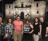 A group of people is posing for a photo at night in front of the historic Alamo mission in San Antonio Texas with one individual holding a Ghost Walk sign suggesting they may be participating in a ghost tour event