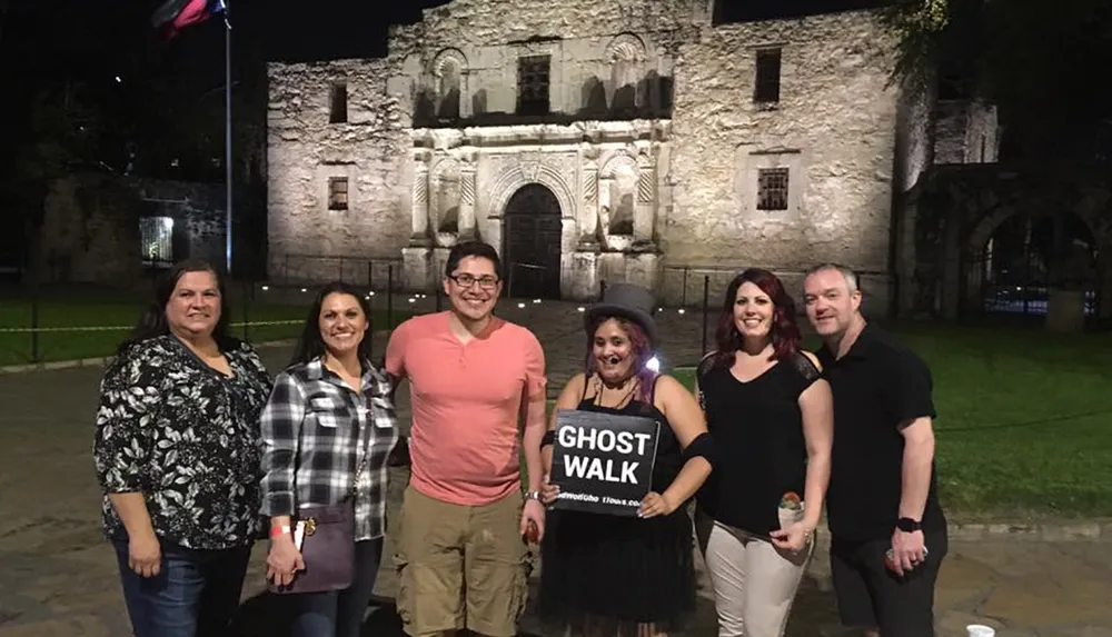 A group of people is posing for a photo at night in front of the historic Alamo mission in San Antonio Texas with one individual holding a Ghost Walk sign suggesting they may be participating in a ghost tour event