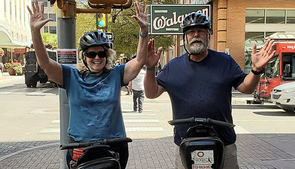 Two people wearing bicycle helmets are joyfully raising their hands in a greeting or celebratory gesture on a city street