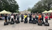 A group of people wearing helmets and riding Segways are waving cheerfully in front of a historic building.