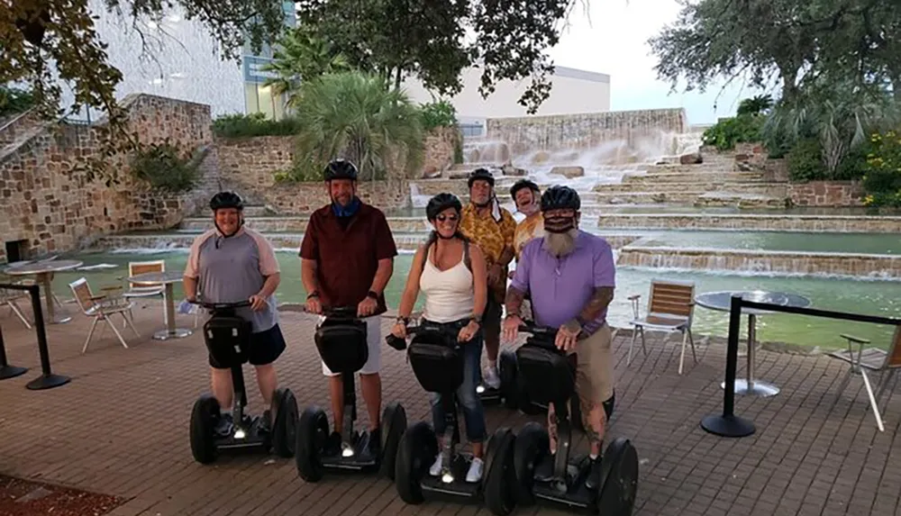 A group of six people poses for a photo on Segways in front of a cascading water feature with most of them wearing helmets and casual attire