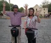 Two happy people are waving while standing on Segways in front of a historical building with other visitors in the background