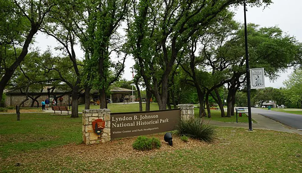 The image shows the entrance to the Lyndon B Johnson National Historical Park with a sign mature trees and visitors in the background