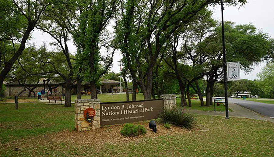 The image shows the entrance to the Lyndon B. Johnson National Historical Park with a sign, mature trees, and visitors in the background.