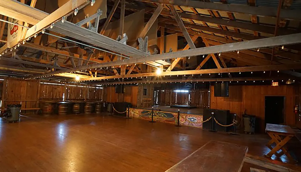 The image shows a spacious wooden-floored hall with a rustic barn-like interior featuring an open beamed ceiling barrels along one wall and a small stage with musical equipment at the far end