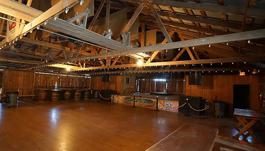 The image shows a spacious, wooden-floored hall with a rustic, barn-like interior, featuring an open beamed ceiling, barrels along one wall, and a small stage with musical equipment at the far end.
