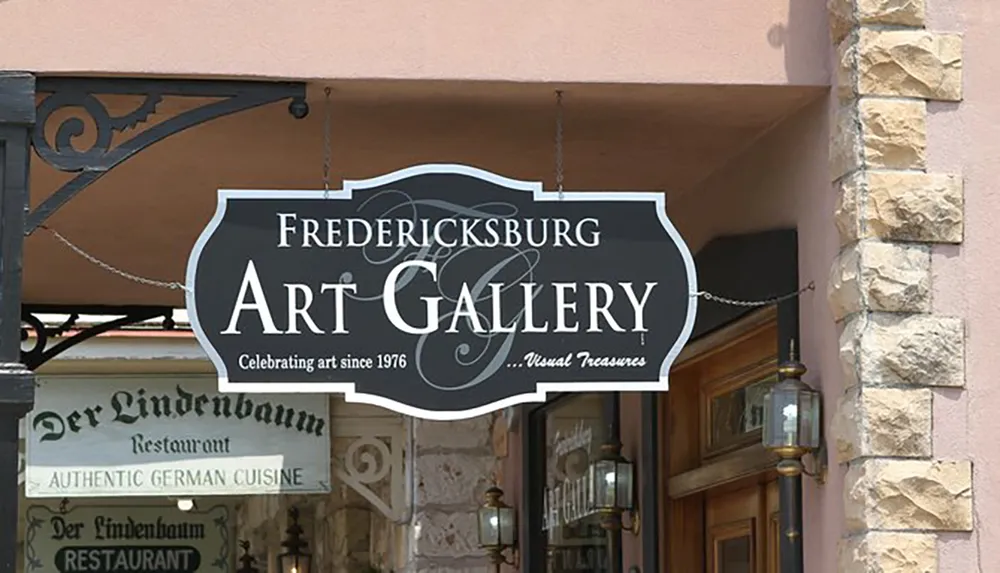 The image shows two hanging signs one for the Fredericksburg Art Gallery with the tagline Celebrating art since 1976 Visual Treasures and another below it for Der Lindenbaum Restaurant advertising Authentic German Cuisine