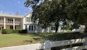 This image shows a traditional two-story white house with a porch and balcony, framed by a white fence and greenery, under a clear sky.