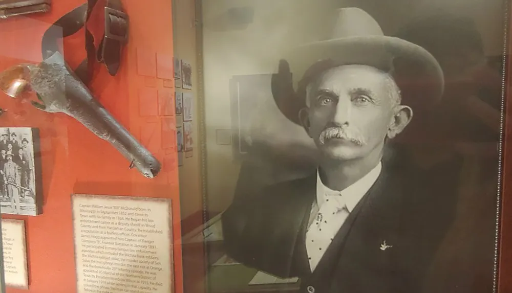 The image shows a museum display featuring historical items including what appears to be a firearm some additional artifacts and the portrait of a mustachioed man wearing a hat likely from the late 19th or early 20th century
