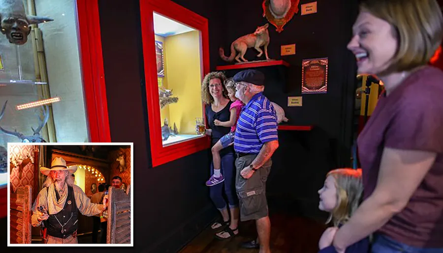 A group of people, including children, are looking at a display containing a bat-like creature in a brightly lit exhibit with an inset image of a man dressed as a cowboy.