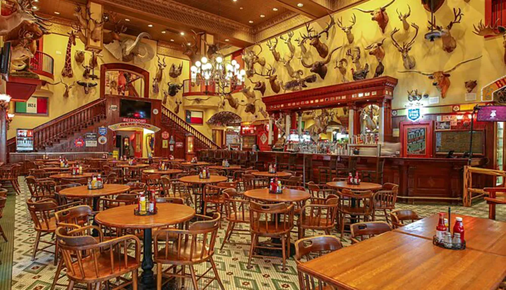 The image depicts a spacious rustic restaurant or bar with numerous wooden tables and chairs and walls adorned with various animal head mounts