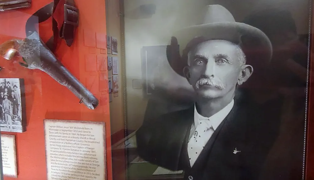 The image shows a display featuring a bespectacled man in a hat alongside a collection of historical artifacts including a gun and photographs