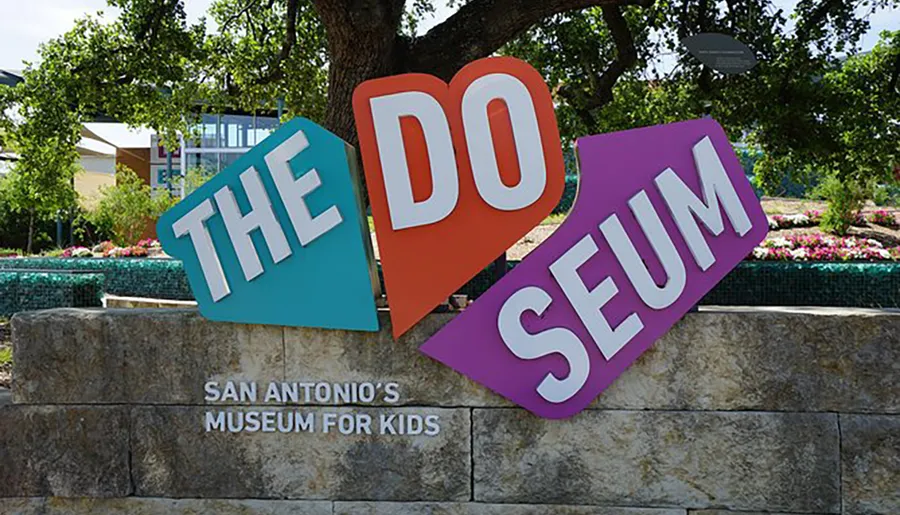 The image displays a colorful, playful sign for The DoSeum, which is San Antonio's museum for kids, positioned against a backdrop of trees, flowers, and a clear sky.