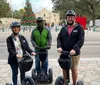 Three smiling people wearing helmets are standing with Segways in front of the Alamo on a sunny day