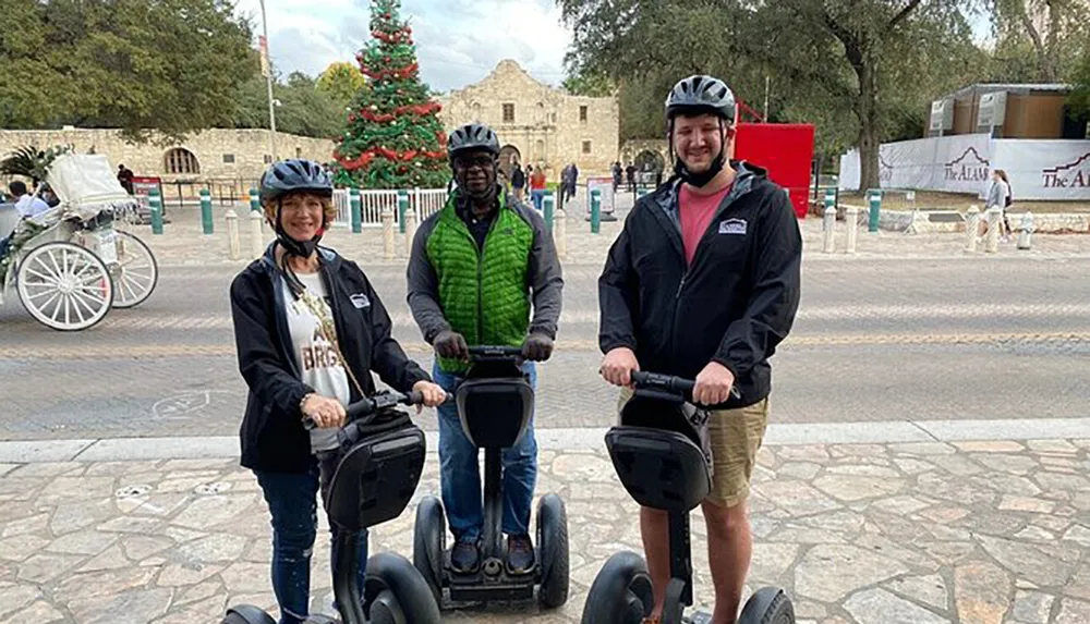 Three people are posing with smiles on their faces while standing on Segways in front of a historic building with a Christmas tree in the background