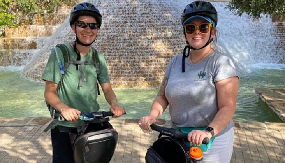 Two people wearing helmets and sunglasses are smiling for a photo while standing next to their Segways in front of a picturesque water fountain