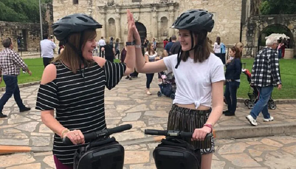 Two people wearing helmets are giving each other a high-five while standing on Segways with a historical building in the background and other visitors walking around