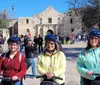 Three smiling people wearing helmets are standing with Segways in front of the Alamo on a sunny day