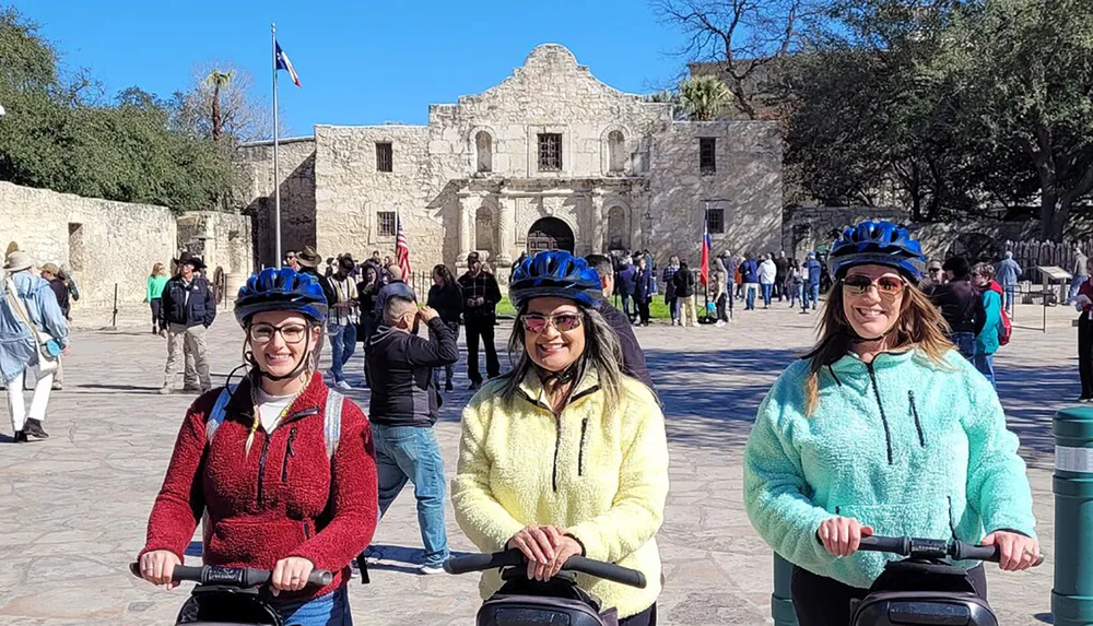 Three smiling people wearing helmets are standing with Segways in front of the Alamo on a sunny day