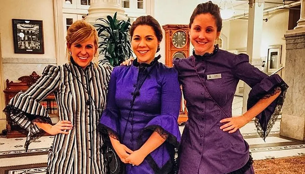 Three smiling women in period-style costumes are posing together in an elegant interior possibly during a themed event or performance