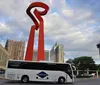 A large red sculptural torch-like structure stands prominently in an urban environment with a tour bus in the foreground and a tower in the distance under a partly cloudy sky