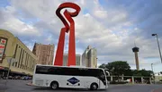 A large red sculptural torch-like structure stands prominently in an urban environment with a tour bus in the foreground and a tower in the distance under a partly cloudy sky.