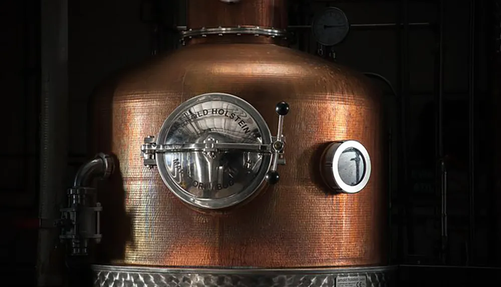 This image shows a close-up of a copper distillation apparatus likely used for producing spirits with a small porthole and various valves gleaming under ambient lighting